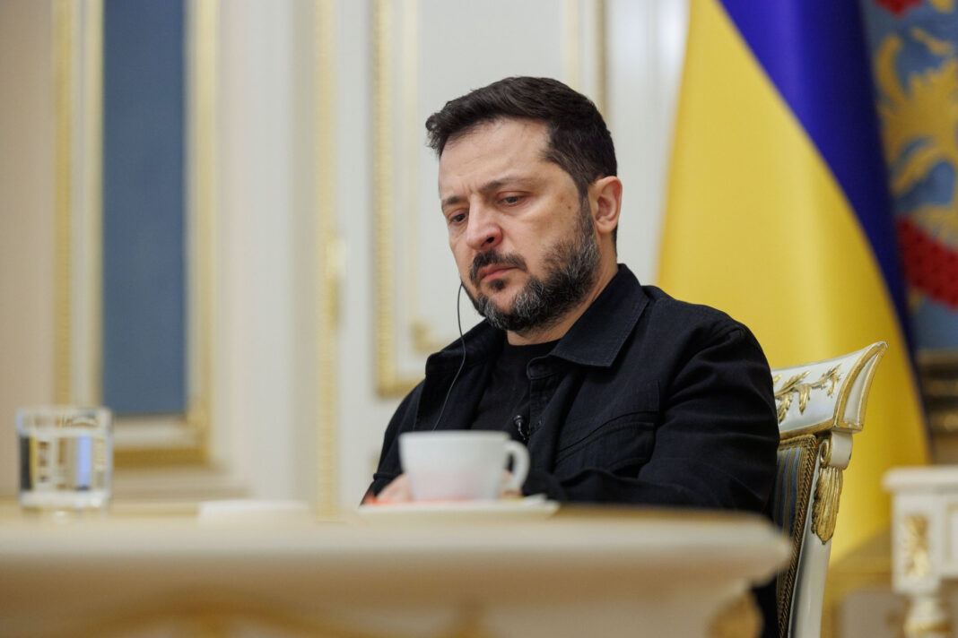 Ukrainian President Volodymyr Zelenskyy sits at a table with a cup and glass of water, a Ukrainian flag behind him, during an interview