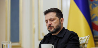 Ukrainian President Volodymyr Zelenskyy sits at a table with a cup and glass of water, a Ukrainian flag behind him, during an interview