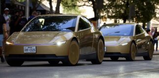 Two gold Tesla Cybercabs drive down a tree-lined street while people on the sidewalk take photos.