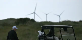 President Donald Trump, wearing a white cap, walks beside a golf cart on a grassy golf course with three wind turbines visible on a hill behind him. (Credit: Forbes/Yahoo News)