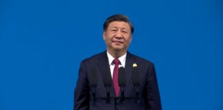 Chinese President Xi Jinping, in a dark suit and red tie, stands behind four microphones against a solid blue background. (File photo : Wikimedia Commons)