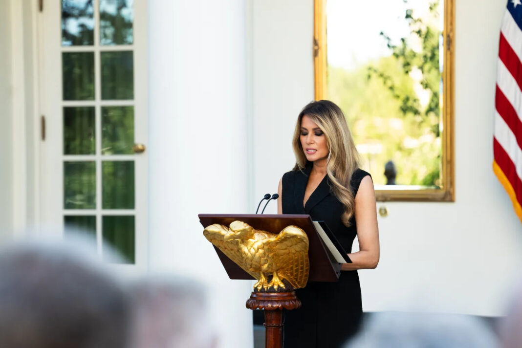First lady Melania Trump, in a black sleeveless dress, reads remarks at a lectern with a gold eagle at the White House Rose Garden.