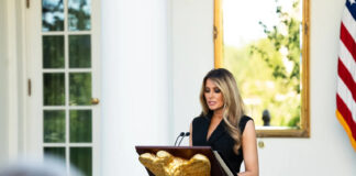 First lady Melania Trump, in a black sleeveless dress, reads remarks at a lectern with a gold eagle at the White House Rose Garden.