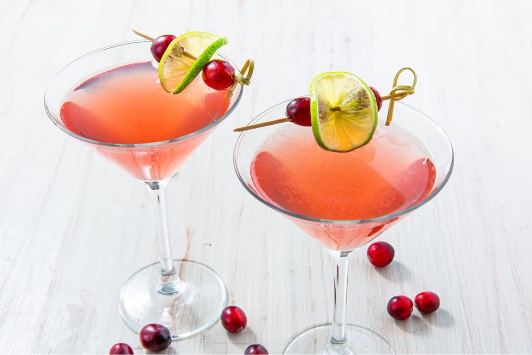 Two pink Cosmopolitan cocktails in martini glasses garnished with lime wheels and cranberries on cocktail picks