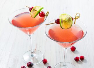 The Cosmopolitan: How to Make the Iconic Pink Cocktail at Home Two pink Cosmopolitan cocktails in martini glasses garnished with lime wheels and cranberries on cocktail picks