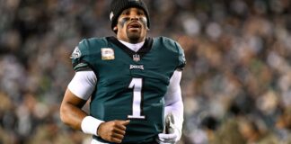 Philadelphia Eagles quarterback Jalen Hurts in his green No. 1 Eagles jersey and a black beanie on the field, with a blurred crowd behind him.