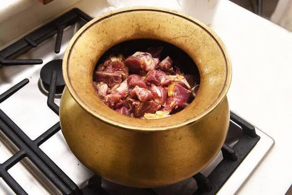 Goat meat in a brass-colored kasaudi on a stovetop before slow cooking