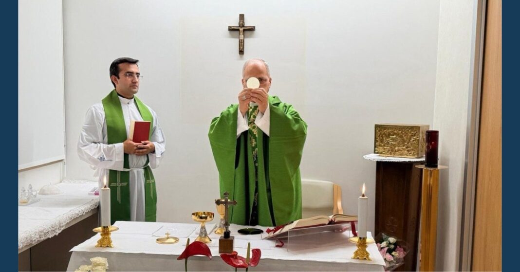 Pope Leo XIV, wearing green vestments, elevates the Eucharistic host during Mass at an altar with lit candles, a chalice and an open book, while an altar server holding a red book stands beside him beneath a wall crucifix.