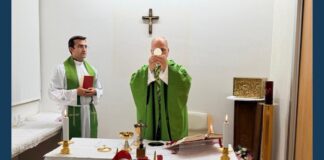 Pope Leo XIV, wearing green vestments, elevates the Eucharistic host during Mass at an altar with lit candles, a chalice and an open book, while an altar server holding a red book stands beside him beneath a wall crucifix.