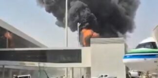 Thick black smoke and flames rise behind a terminal building at King Khalid International Airport in Riyadh, Saudi Arabia, as a passenger jet sits parked nearby