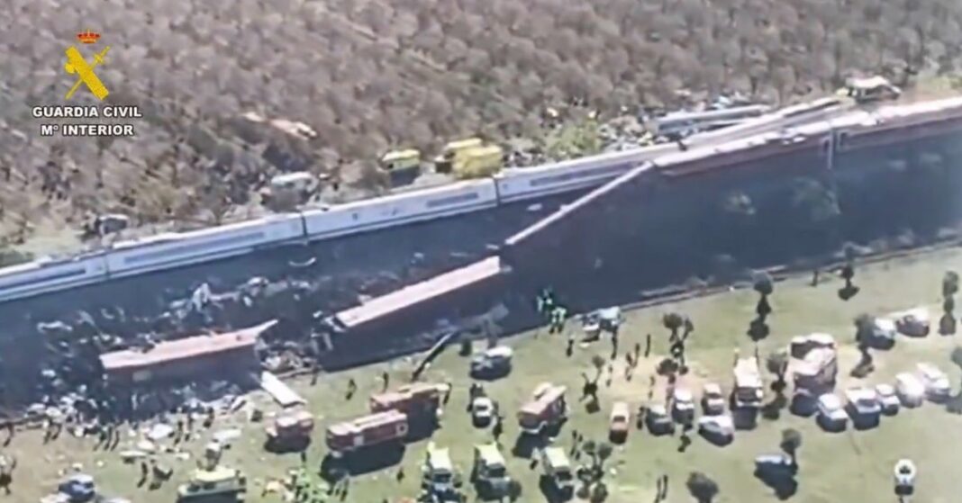 Aerial view of damaged train carriages beside railway tracks, with emergency vehicles and responders nearby.