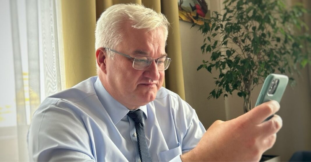 Andrii Sybiha wearing glasses and a tie looks at a smartphone beside a window.