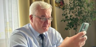 Andrii Sybiha wearing glasses and a tie looks at a smartphone beside a window.