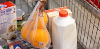 A shopper places a bag of oranges in a shopping cart next to a gallon of milk and other groceries